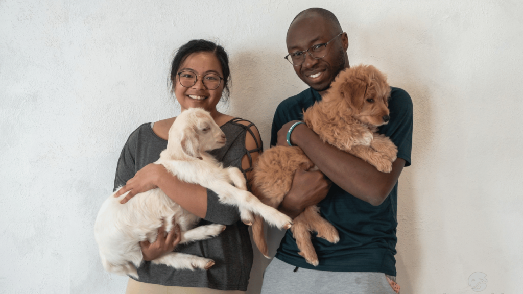 Smiling man and woman holding a baby goat and a fluffy puppy, representing the joy of gifting a Karmably experience voucher.
